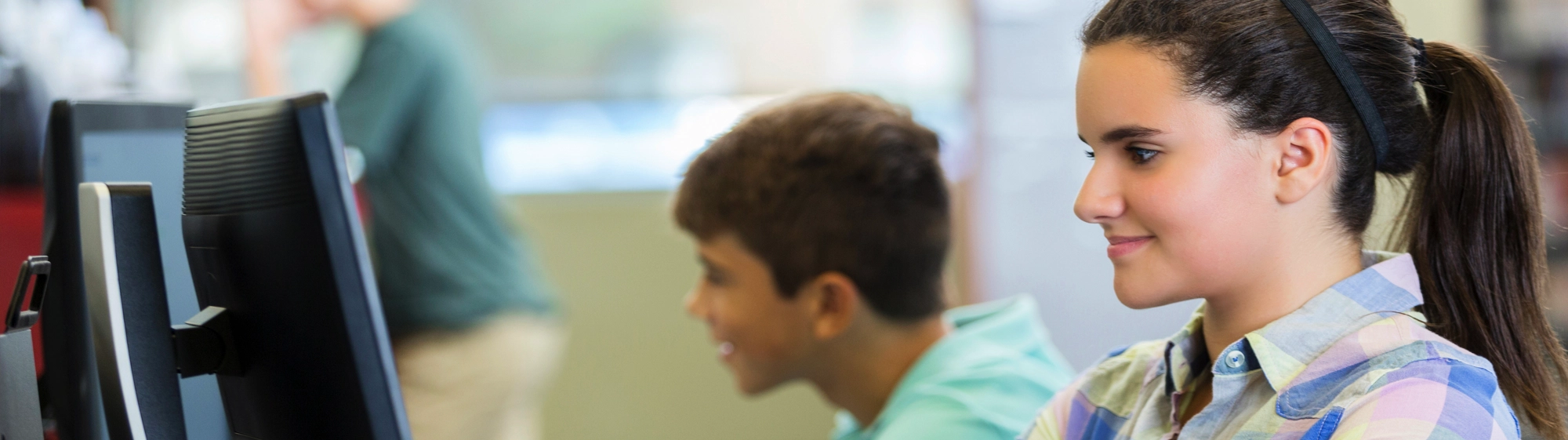 High Schooler on computer, AFAB is looking at screen, smiling, wearing a headband and a plaid shirt for Varied Practice Reading