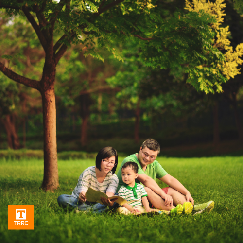 Family sitting on the grass by a tree, reading a book together.