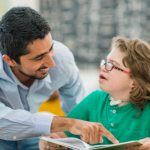A male teacher pointing to a book that a student is holding in an open hand, the student is looking up at the teacher.