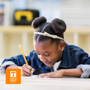 Student seated at a desk writing with a pencil on a Writing Mat from the TRRC, student is smiling, her hair is in two buns, and she's wearing a jean jacket