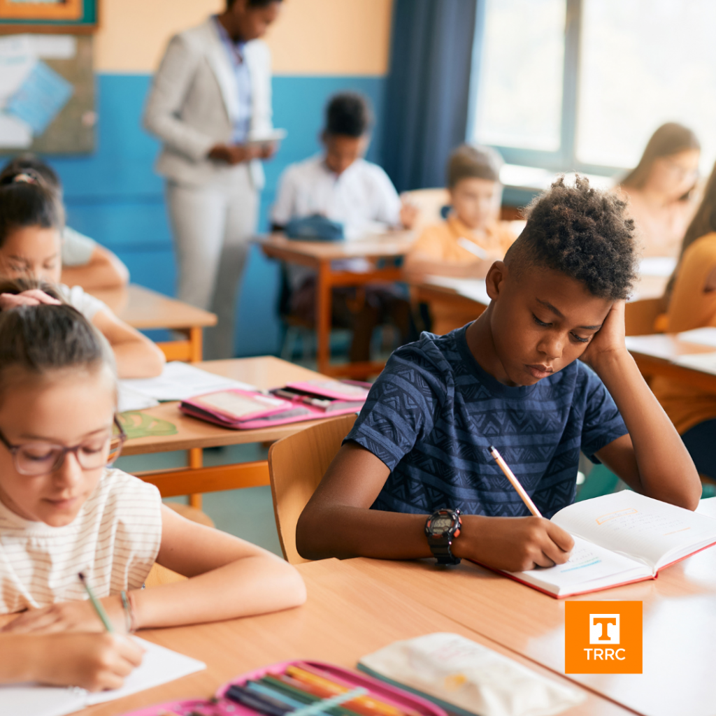 A classroom of students writing, one student is zoomed in, writing in a book, the teacher is in the back of the classroom