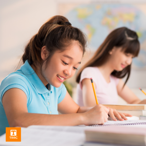 an upper elementary girl and another female student are writing in a paper notebook at a desk in a classroom